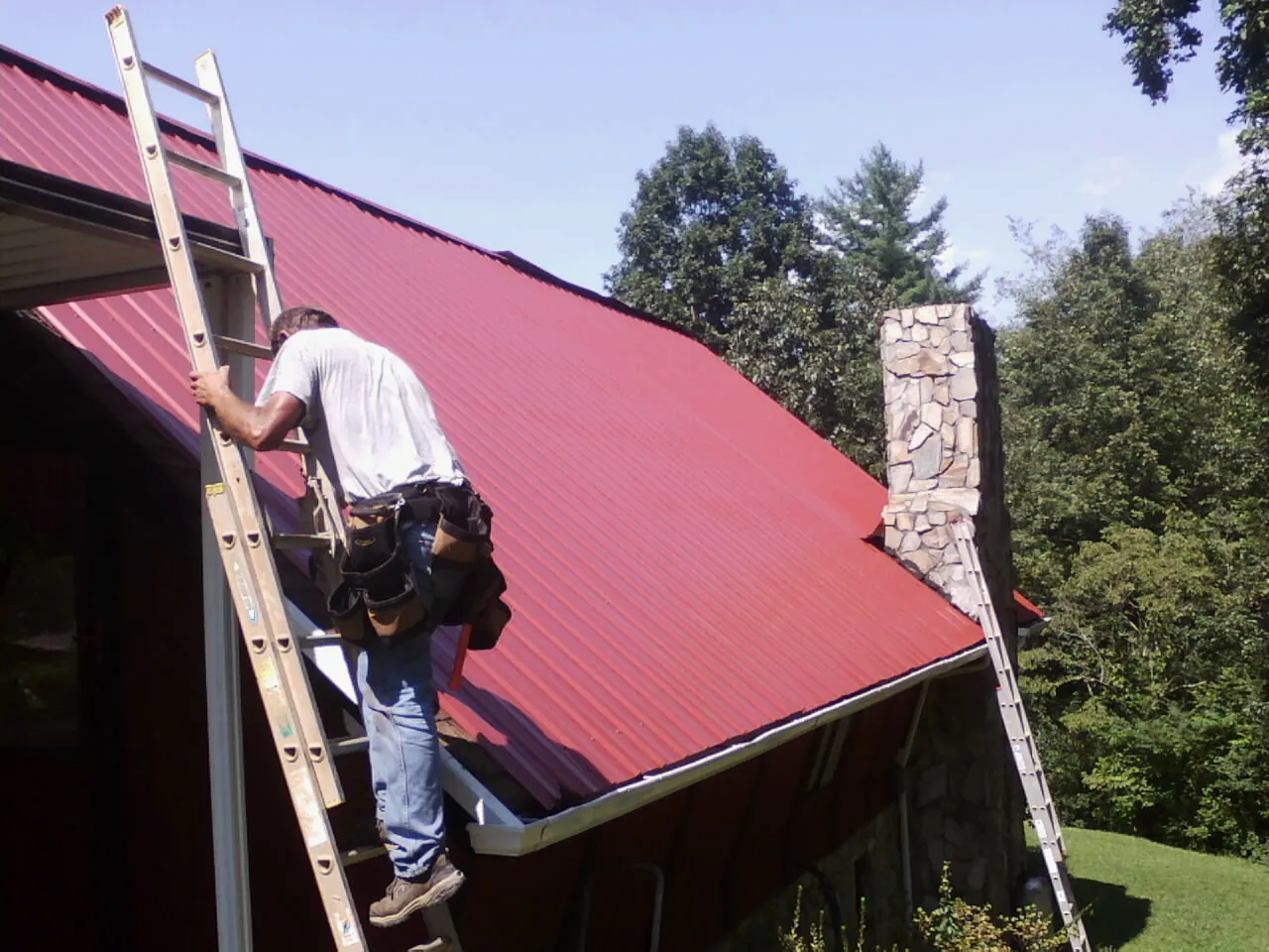 Roofer on a ladder beside a red metal roof and stone chimney — Soffit & Fascia Repair by Roofing Brothers in Santa Cruz